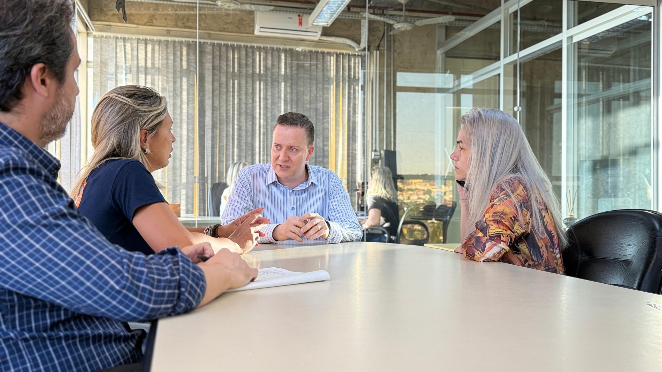 A imagem mostra quatro pessoas sentadas em torno de uma mesa de conferências, aparentemente em uma reunião de negócios. Vamos analisar os detalhes: * *Ambiente:* O local parece ser um escritório moderno, com uma grande janela de vidro que oferece uma vista externa. O espaço é bem iluminado e possui divisórias de vidro, sugerindo um ambiente de trabalho colaborativo e aberto. * *Participantes:* Há quatro indivíduos presentes: * *Homem à esquerda (de camisa xadrez azul):* Ele parece ser um dos principais participantes da reunião, possivelmente um líder ou tomador de decisão, dado sua posição e a forma como ele está envolvido na conversa. * *Mulher com cabelo loiro (à direita do homem de camisa xadrez):* Ela parece estar ativamente participando da discussão, interagindo com o homem de camisa azul e o homem sentado em frente. Sua postura sugere engajamento e atenção ao que está sendo discutido. * *Homem de camisa listrada (ao centro):* Ele está sentado de frente para as outras duas pessoas, e sua expressão e postura sugerem que ele está apresentando algo ou respondendo a perguntas. Ele parece ser um ponto focal da conversa. * *Mulher com cabelo grisalho longo (à direita, de costas para a câmera):* Ela parece estar menos envolvida na discussão principal, possivelmente ouvindo ou aguardando sua vez de falar. Ela está ao telefone ou com um dispositivo semelhante. * *Dinâmica da reunião:* A imagem sugere uma dinâmica de discussão ou brainstorming. A proximidade dos participantes, suas expressões faciais e a posição de suas mãos indicam que estão envolvidos em uma troca de ideias ativa. O homem de camisa listrada parece ser o centro da discussão, possivelmente respondendo a perguntas ou apresentando informações. * *Elementos adicionais:* Há um caderno ou bloco de notas na mesa, sugerindo que anotações estão sendo feitas durante a reunião. Em resumo, a imagem captura um momento de uma reunião de negócios em um ambiente de trabalho moderno. A dinâmica da reunião parece ser colaborativa e focada em uma discussão específica, com o homem de camisa listrada como um ponto central de informação. A mulher de cabelo grisalho parece estar em uma ligação separada, sugerindo uma possível multitarefa ou uma conversa paralela.