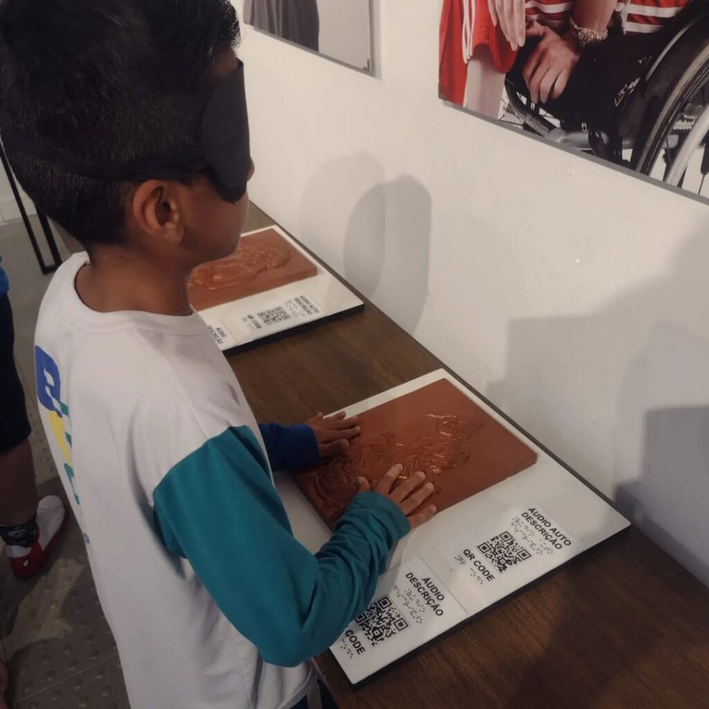 Criança com os olhos vendados toca uma prancha tátil em uma mesa expositora. A prancha contém relevo artístico e um QR Code para audiodescrição. A criança veste camiseta branca com mangas azuis e está concentrada na atividade. Ao fundo, vê-se parte de uma imagem fotográfica exposta na parede, com destaque para uma pessoa em cadeira de rodas. A cena faz parte de uma atividade educativa inclusiva na Pinacoteca.