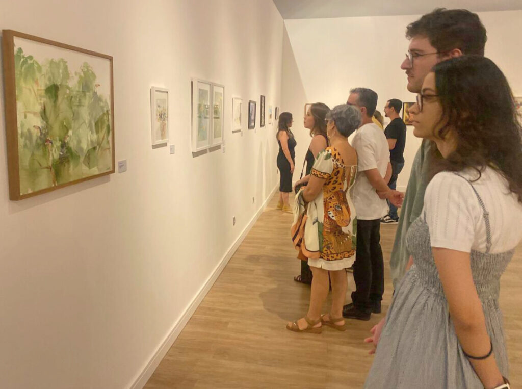 Visitantes de perfil observam com atenção uma série de quadros alinhados em uma parede branca. As obras apresentam paisagens e elementos da natureza. O piso é de madeira clara.
