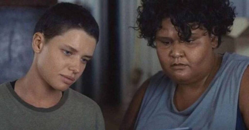 Cena do filme Cidade Campo mostra duas mulheres em ambiente interno com pouca luz. À esquerda, uma mulher de cabelo raspado e expressão séria olha para baixo; à direita, uma mulher de cabelo cacheado curto e volumoso, com expressão de concentração, também olha para baixo. As duas estão lado a lado, com rostos próximos, lendo ou observando algo fora do quadro.