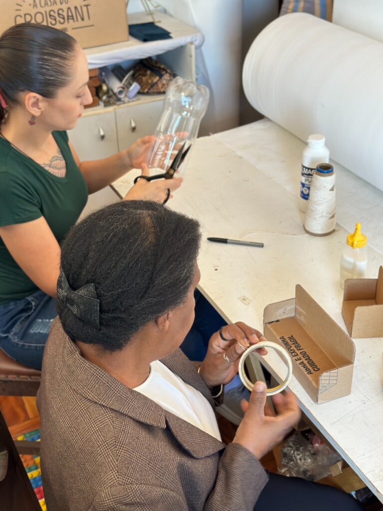 A imagem mostra duas mulheres sentadas em uma mesa, aparentemente em um ateliê ou sala de artesanato. Uma mulher mais jovem está cortando uma garrafa de plástico com uma tesoura, enquanto uma mulher mais velha está usando fita adesiva em uma pequena caixa de papelão. Há vários materiais de artesanato na mesa, incluindo cola, barbante e um rolo de tecido. O ambiente parece ser acolhedor e criativo, sugerindo um projeto de artesanato colaborativo. As mulheres parecem concentradas e envolvidas em seu trabalho. A imagem é bem iluminada e nítida, mostrando os detalhes dos materiais e das ações das mulheres.