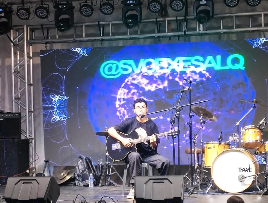 A imagem mostra um jovem sentado num banco, tocando violão num palco. Ele usa óculos e uma camiseta escura. Ao fundo, há uma tela grande com um design abstrato e o texto "@SVQEXESALQ". Há também uma bateria e equipamento de som no palco. O ambiente parece ser um evento ao ar livre ou num grande salão de eventos. A iluminação é profissional, com holofotes acima do palco. A imagem é nítida e bem iluminada, capturando bem a atmosfera do evento.