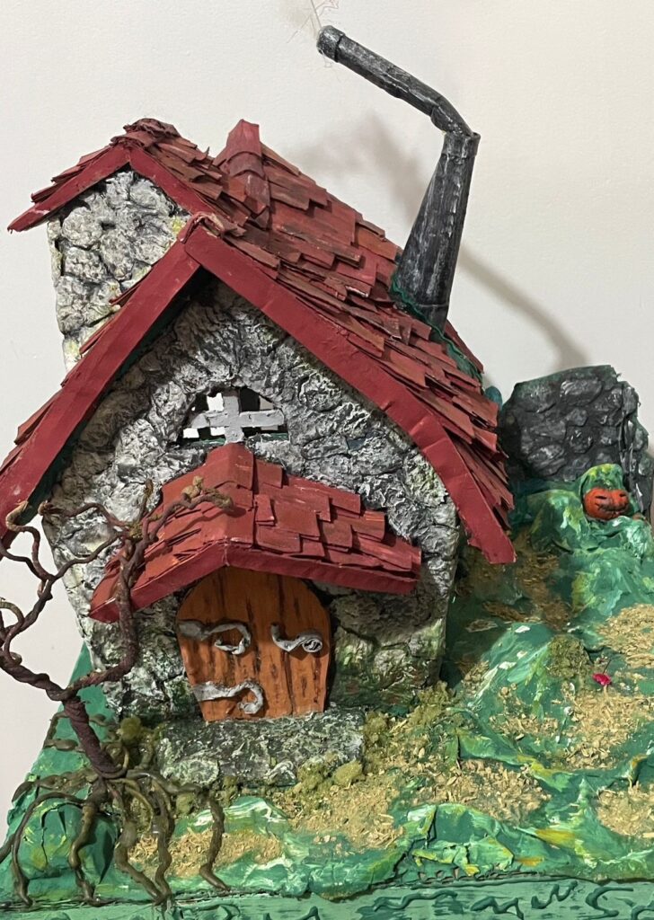 A imagem mostra uma maquete detalhada de uma pequena casa de pedra com telhado vermelho e uma chaminé. A casa parece velha e desgastada, com alguns danos visíveis no telhado e nas paredes. A porta é de madeira e parece antiga, com detalhes esculpidos. A casa está situada em uma paisagem verdejante, com pequenas árvores e arbustos ao redor. O cenário inclui uma pequena abóbora esculpida, adicionando um toque de Halloween. A maquete parece ser feita de papel machê ou material semelhante, cuidadosamente pintado e texturizado para dar a impressão de pedra e madeira. O nível de detalhe é impressionante, com texturas realistas e cores vibrantes. A composição geral é criativa e bem executada, evocando uma atmosfera encantadora e um tanto sinistra.