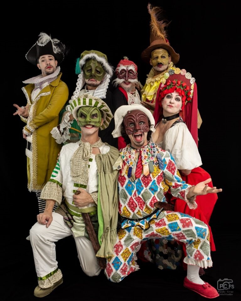 A imagem mostra um grupo de sete atores usando fantasias e máscaras elaboradas, posados contra um fundo preto. As fantasias são vibrantes e detalhadas, com uma variedade de cores e texturas, sugerindo um contexto teatral ou de performance. As máscaras são expressivas e contribuem para a atmosfera dramática da cena. Os atores parecem representar personagens distintos, cada um com sua própria individualidade, expressa através da vestimenta e postura. A imagem é bem iluminada, com foco nítido nos atores, destacando os detalhes das fantasias e máscaras. A composição é dinâmica, com os atores posicionados de forma a criar uma sensação de movimento e interação entre eles. O estilo geral sugere uma peça de teatro, possivelmente com temática histórica ou de comédia.