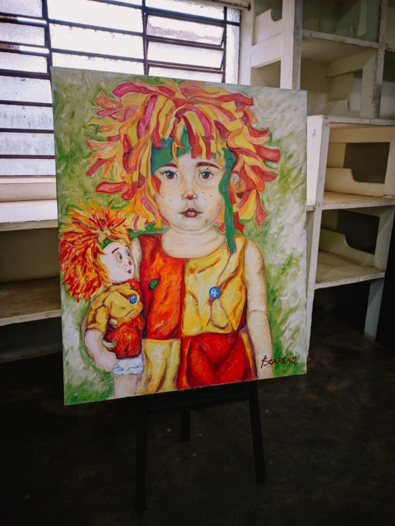 A imagem mostra uma pintura a óleo vibrante e expressiva de uma criança com uma grande peruca colorida, segurando um boneco de aparência semelhante. A criança está vestida com um traje multicolorido, principalmente em tons de vermelho, amarelo e laranja, que combinam com a peruca. O estilo da pintura é expressivo, com pinceladas visíveis e cores saturadas. O fundo é um verde suave e desfocado, que contrasta com as cores vibrantes da figura principal. A pintura está em um cavalete, e o ambiente parece ser um estúdio ou galeria de arte, com prateleiras de madeira visíveis ao fundo. A assinatura do artista, "Scarenzi," está visível no canto inferior direito da pintura. A imagem como um todo transmite uma sensação de alegria e criatividade.