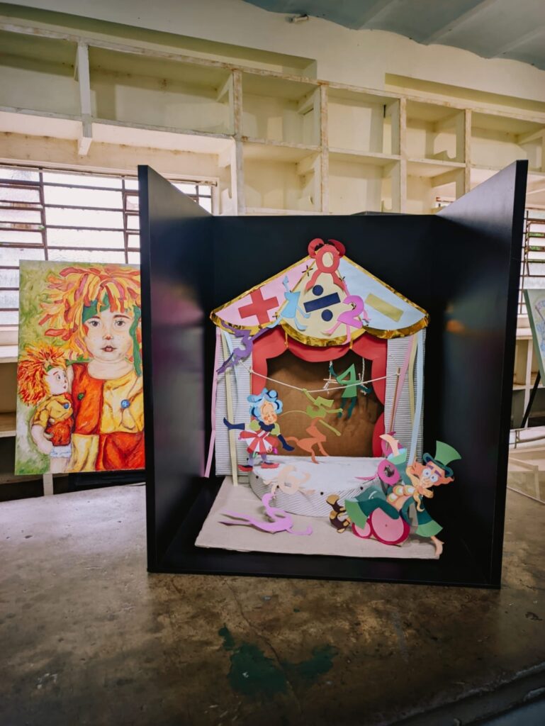 A imagem mostra uma instalação artística dentro de um espaço que parece ser um estúdio ou ateliê. Há uma caixa preta que contém uma maquete de um palco de teatro de fantoches, com fantoches de papel coloridos representando diferentes personagens. A maquete é rica em detalhes, com um telão, cortinas e vários elementos decorativos. Ao lado da caixa, há uma pintura a óleo em tela representando uma menina com cabelo vermelho e uma boneca. O fundo mostra prateleiras de madeira, janelas e paredes desgastadas pelo tempo, sugerindo um ambiente antigo e criativo. O chão é de concreto, com marcas de uso e desgaste. A iluminação é suave e natural, provavelmente proveniente de uma janela próxima. A cena transmite uma atmosfera lúdica e artística.