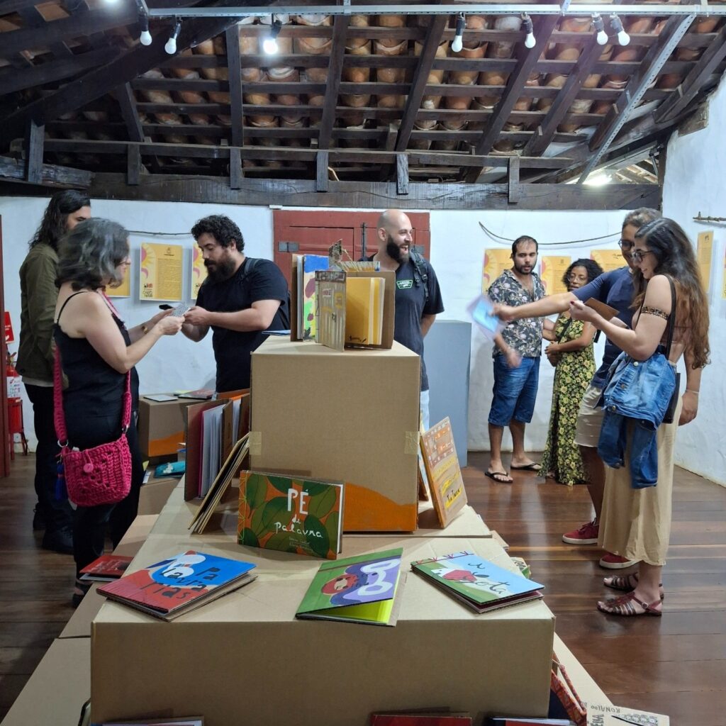 A imagem mostra uma exibição de livros ilustrados numa sala de paredes brancas e telhado de madeira exposto com vigas de madeira e telhas de barro. Várias pessoas estão a olhar e a interagir com os livros, que estão dispostos em caixas de cartão. Os livros parecem ser feitos à mão e têm capas vibrantes e coloridas. As pessoas na imagem são de diversas idades e etnias, e parecem estar a desfrutar da exposição. O ambiente é casual e acolhedor. A iluminação é suave e natural, e a imagem no geral tem uma atmosfera calma e convidativa.