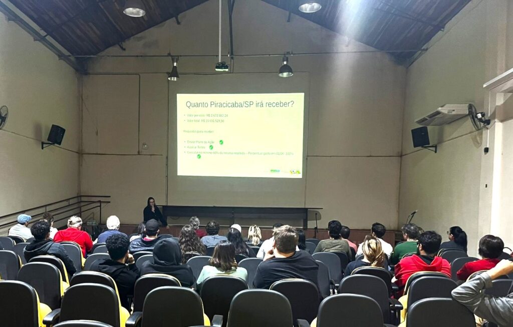 A imagem mostra uma apresentação em uma sala de conferências ou auditório. Um projetor exibe uma apresentação em português sobre quanto Piracicaba, São Paulo, receberá em financiamento. A apresentação lista os requisitos para receber os fundos, incluindo o envio de um plano de ação e a assinatura de um termo. A sala está repleta de pessoas sentadas em cadeiras individuais, presumindo-se que sejam participantes da apresentação. Uma mulher está de pé na frente da tela, possivelmente a palestrante. A atmosfera parece ser formal e voltada para negócios. A iluminação é discreta, e o ambiente parece ser um salão ou espaço de eventos adaptado para palestras.