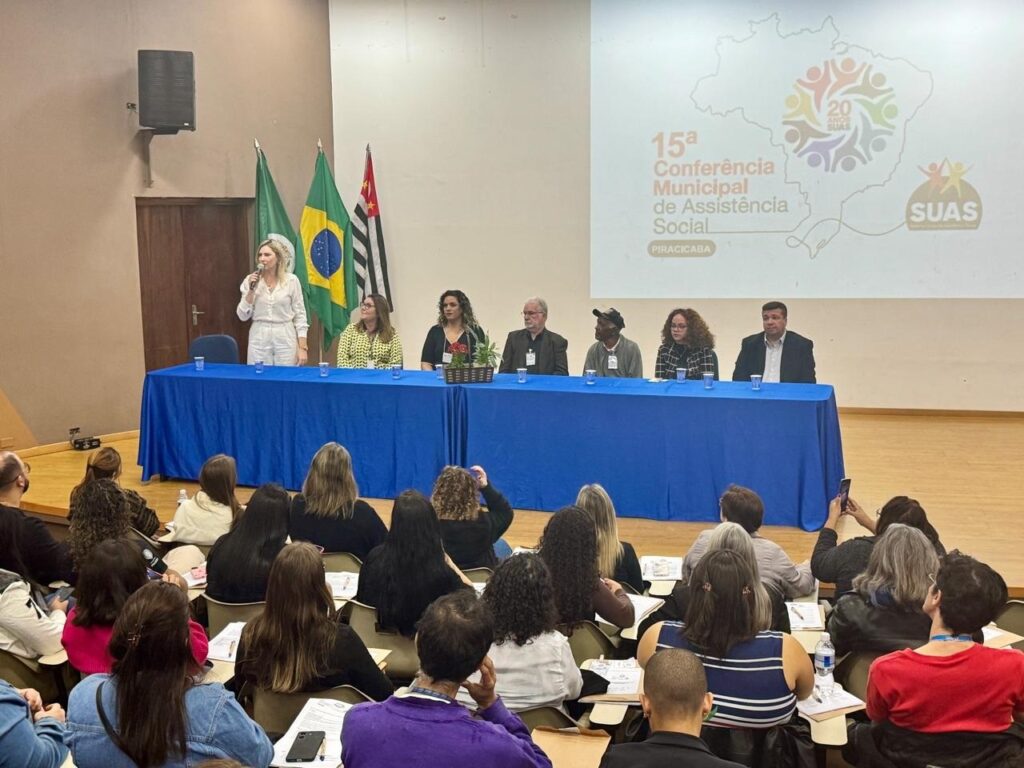 A imagem mostra uma conferência em andamento, aparentemente sobre assistência social, em Piracicaba. Uma mulher está no palco, usando um microfone e falando para a plateia. No palco, também há uma mesa com outras cinco pessoas sentadas, aparentemente um painel. Há bandeiras brasileiras e de outros estados atrás deles. A apresentação inclui um grande banner indicando a "15ª Conferência Municipal de Assistência Social", com o logo do SUAS (Sistema Único de Assistência Social). A plateia é composta por muitas pessoas sentadas em cadeiras, prestando atenção. O ambiente parece ser uma sala de conferências ou auditório. A atmosfera é formal e atenta.
