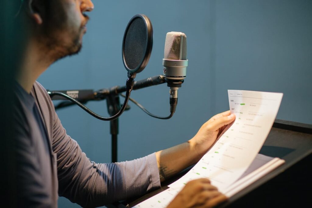 A imagem mostra um homem sentado em frente a um microfone, lendo um roteiro. Ele está usando uma camiseta cinza e parece estar em um estúdio de gravação. O microfone é um microfone condensador com um pop filter, e o roteiro está em uma superfície inclinada. A iluminação é suave e o fundo é desfocado. O homem parece estar concentrado em sua leitura. A imagem é bem composta e a qualidade é alta.