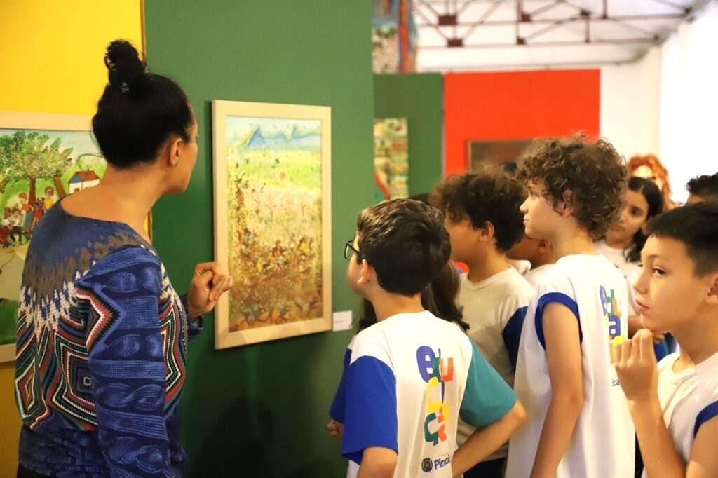 A imagem mostra um grupo de crianças em idade escolar em uma galeria de arte, observando atentamente uma pintura. Uma mulher, provavelmente uma professora ou guia, está ao lado deles, apontando para a obra de arte e explicando-a. As crianças parecem engajadas e curiosas, com expressões focadas em seus rostos. Elas vestem uniformes escolares, sugerindo uma visita de campo organizada. O fundo mostra outras pinturas na parede e a arquitetura da galeria. A iluminação é suave e natural, criando um ambiente acolhedor e educativo. A cena captura um momento de aprendizado e apreciação da arte, mostrando a importância da educação artística para jovens. A imagem é vibrante e bem composta, com cores e detalhes que transmitem uma atmosfera animada e envolvente.