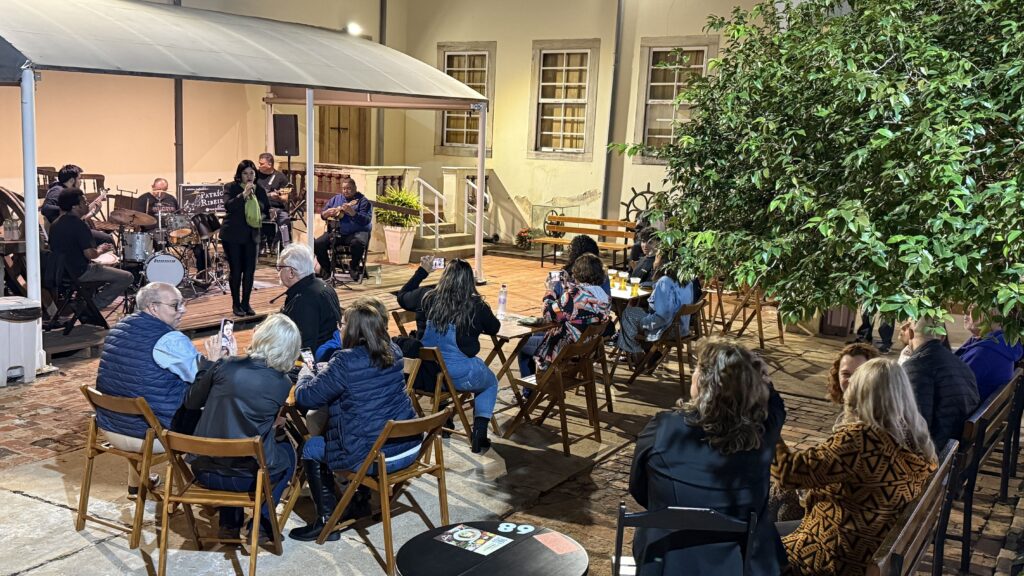 A imagem mostra uma apresentação musical ao ar livre em um pátio. Uma banda está tocando em um palco improvisado sob uma tenda, enquanto um público sentado em mesas e cadeiras assiste. O local parece ser um pátio ou jardim, com árvores e bancos visíveis. O ambiente é aconchegante e informal, com iluminação suave que sugere uma atmosfera noturna. As pessoas na plateia parecem relaxadas e estão interagindo entre si, algumas fotografando a apresentação. A imagem captura um momento de entretenimento ao ar livre em um ambiente agradável.