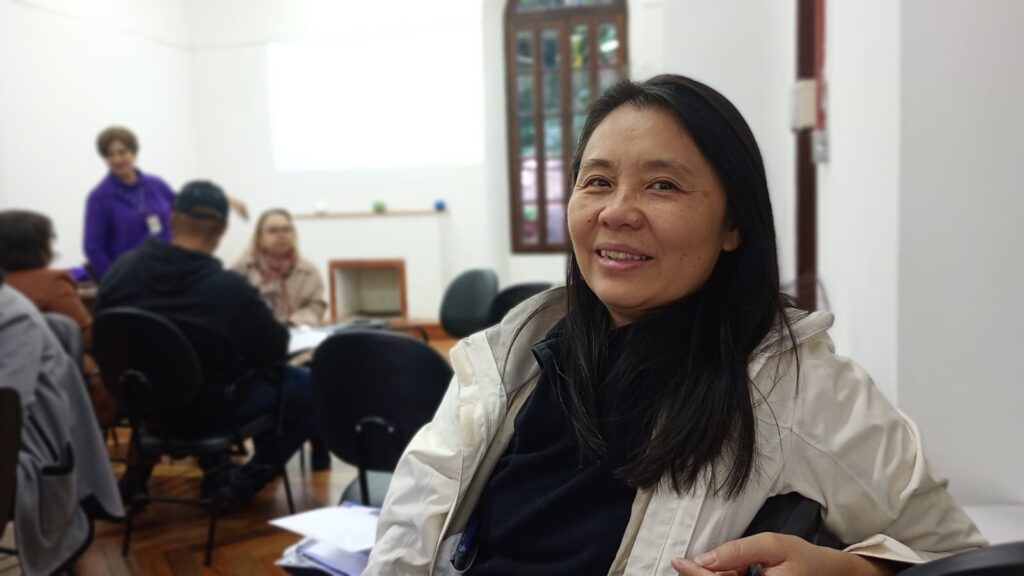 A imagem mostra uma mulher de meia-idade com cabelos pretos e longos, sorrindo para a câmera. Ela está usando uma jaqueta bege clara e uma blusa escura por baixo. Ela parece estar sentada em uma cadeira em uma sala de aula ou sala de conferências, com outras pessoas sentadas em segundo plano, levemente fora de foco. As outras pessoas parecem estar em uma reunião ou apresentação. O fundo é uma sala simples e bem iluminada com paredes brancas, uma janela e uma lareira. A mulher está no foco principal da imagem, e sua expressão facial é amigável e acolhedora. A atmosfera geral da imagem é calma e profissional.