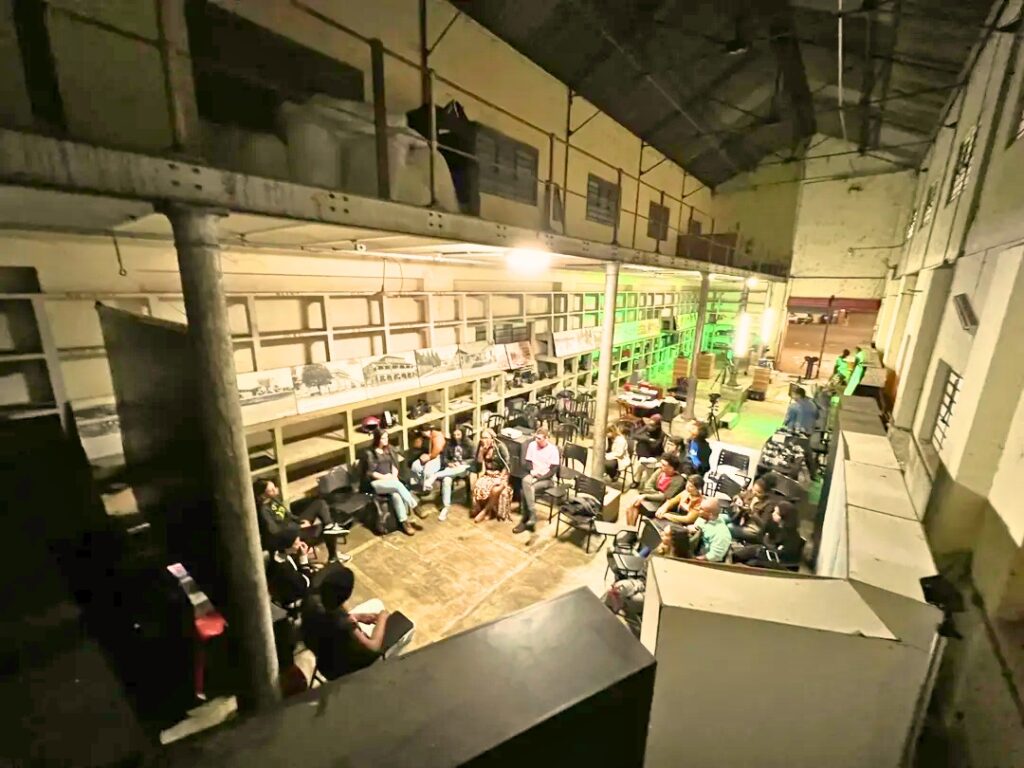 A imagem mostra um amplo espaço interior, possivelmente um armazém ou fábrica adaptado, onde um grupo de pessoas está sentado em cadeiras, aparentemente assistindo a um evento ou apresentação. O espaço é amplo e alto, com uma estrutura de metal elevada que suporta um mezanino ou plataforma superior. Há prateleiras ao longo das paredes, algumas contendo objetos ou itens arquivados. A iluminação é baixa e difusa, criando uma atmosfera mais intimista. As pessoas estão sentadas em grupos, algumas conversando, outras prestando atenção em algo no centro do espaço. A arquitetura do local sugere um edifício antigo, com paredes de tijolo aparente e vigas de madeira expostas. O chão parece ser de concreto. No geral, a imagem transmite uma sensação de evento comunitário ou reunião informal num espaço industrial reconvertido.