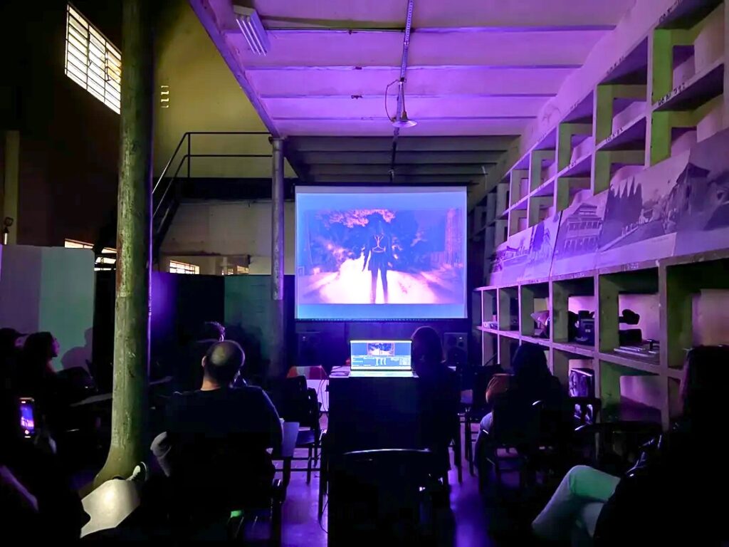 A imagem mostra uma sala escura, possivelmente um estúdio de edição de vídeo ou um espaço de exibição, com um projetor exibindo uma cena de filme na parede. A cena parece ser de um filme de ficção científica ou fantasia, mostrando uma figura caminhando em um ambiente desértico ou futurista. Há várias pessoas sentadas em cadeiras, assistindo ao filme. A iluminação é baixa e difusa, com uma tonalidade púrpura dominante, criando uma atmosfera sombria e concentrada. No centro da sala, há uma mesa com um computador, provavelmente usado para edição de vídeo, com um teclado e uma tela visíveis. Nas laterais da sala, existem prateleiras cheias de equipamentos de produção de vídeo e outros objetos, sugerindo que o local é usado para edição e pós-produção de filmes. A arquitetura do espaço é antiga e industrial, com vigas de madeira expostas no teto e colunas de concreto. A atmosfera geral é de um ambiente de trabalho criativo e intenso, focado na produção de filmes.