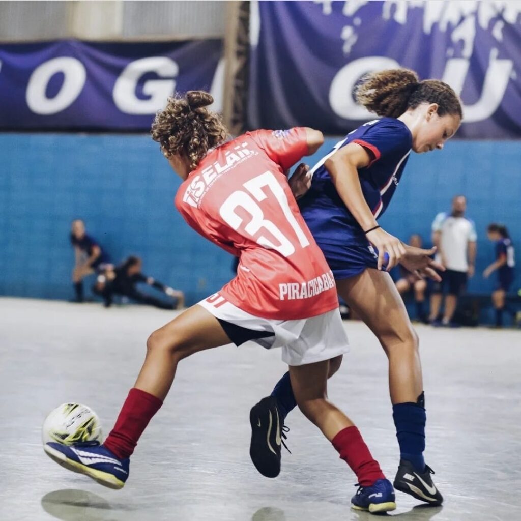 A imagem mostra duas jovens jogadoras de futebol de salão em plena ação durante uma partida. Uma delas, vestindo uma camisa vermelha com o número 37 e o nome "PIRACICABA", está driblando a outra jogadora, que veste uma camisa azul escura. Ambas as jogadoras estão usando meias e chuteiras, e a ação é intensa, com muita dinâmica e movimento capturados no instante. O fundo mostra uma quadra de esportes indoor, com uma faixa azul com letras brancas e algumas pessoas observando a partida no fundo, ligeiramente desfocadas, enfatizando a ação principal entre as duas jogadoras. A iluminação é boa, com detalhes visíveis nas roupas e expressões faciais das atletas. A imagem transmite a energia e competição de um jogo de futebol de salão, com foco na habilidade e destreza das jogadoras em ação.