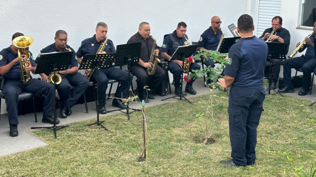 A imagem mostra uma banda de músicos uniformizados, possivelmente uma banda militar ou policial, tocando instrumentos de sopro ao ar livre. Eles estão em formação, com um maestro à frente conduzindo a apresentação. Os músicos estão sentados em cadeiras, com seus instrumentos de sopro, incluindo saxofones e trompetes, posicionados em frente a eles. O cenário é um jardim gramado, com arbustos e árvores ao fundo. A atmosfera é formal e tranquila, sugestiva de uma apresentação ou ensaio musical. Os uniformes são escuros e os instrumentos brilhantes, criando um contraste visual. A imagem é bem iluminada, indicando que a foto foi tirada em um dia ensolarado.