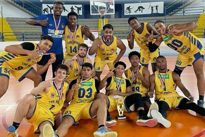 A imagem mostra uma equipe de basquete vestindo uniformes amarelos e azuis. Os jogadores estão sentados e em pé em uma quadra de basquete, sorrindo para a câmera. Eles estão segurando medalhas de ouro e um troféu. A maioria dos jogadores está usando shorts e regatas amarelas com o logotipo "The Ring Basketball" e números em seus uniformes. Alguns jogadores estão usando medalhas de ouro em volta do pescoço. O fundo mostra uma quadra de basquete interna com um quadro de basquete e um mural na parede. A iluminação é brilhante e a imagem é nítida e clara. A atmosfera geral da imagem é alegre e festiva, sugerindo uma celebração de uma vitória ou conquista.