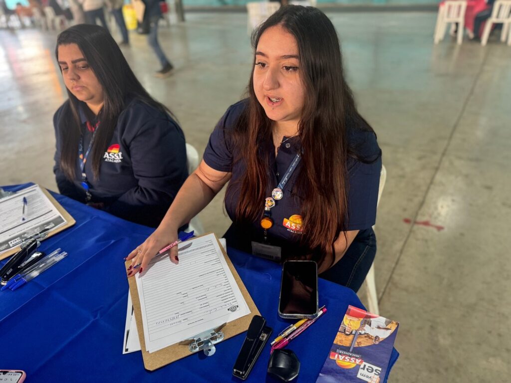 A imagem mostra duas mulheres sentadas a uma mesa, aparentemente em um evento ou feira. Uma delas está de uniforme azul-escuro com o logotipo da "ASSAI ATACADISTA", e parece estar preenchendo uma documentação ou formulário em uma prancheta. A outra mulher está sentada ao seu lado, também vestindo um uniforme semelhante. Há diversos objetos sobre a mesa, incluindo canetas, uma grampeadora, um celular e um panfleto ou catálogo da ASSAI ATACADISTA. O ambiente parece ser um espaço amplo e aberto, com piso de concreto. A mulher que está preenchendo o formulário parece estar interagindo com alguém fora do quadro, possivelmente um cliente ou visitante do evento. A atmosfera parece ser formal e profissional.