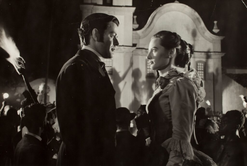 A imagem é uma fotografia em preto e branco de um casal vestido com roupas de época, possivelmente de um filme ou peça de teatro. Eles estão em pé, um de frente para o outro, em um ambiente externo à noite, iluminados por tochas, com outras pessoas ao fundo. O homem está vestido com um casaco escuro e uma gravata, enquanto a mulher veste um vestido elaborado com mangas compridas e babados. As expressões faciais deles sugerem um momento íntimo ou romântico. O fundo mostra uma construção arquitetônica de estilo colonial, com sombras projetadas na parede, possivelmente de pessoas ou árvores. A cena parece ser um evento social ou festivo, considerando as pessoas e as tochas em segundo plano. A qualidade da imagem sugere uma fotografia antiga, talvez de uma produção cinematográfica clássica. A composição da imagem coloca o foco no casal, enquanto o fundo fornece contexto e atmosfera. O contraste entre a luz e a sombra cria uma atmosfera dramática e romântica.