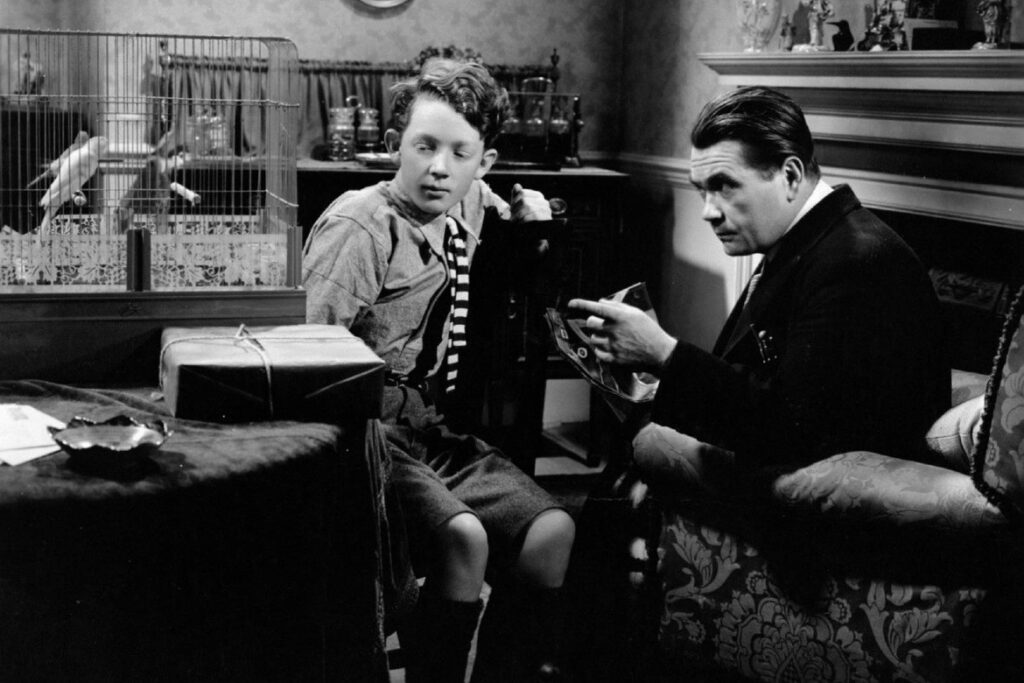 A imagem em preto e branco mostra um menino e um homem sentados em uma sala. O menino, que parece ter cerca de dez anos, está sentado em uma cadeira à esquerda do quadro, usando uma camisa de botão, gravata listrada e calças curtas. Ele parece estar observando algo que o homem está segurando. O homem, que parece ter cerca de quarenta anos, está sentado em uma poltrona à direita do quadro, usando um terno. Ele parece estar mostrando algo ao menino. Há uma gaiola de pássaros ao fundo, e uma caixa embrulhada em papel pardo está sobre uma mesa entre eles. A sala parece ser uma sala de estar, com uma lareira e alguns móveis visíveis. A iluminação da imagem é suave e difusa, criando uma atmosfera calma e íntima. A imagem como um todo parece ser uma cena de um filme ou peça de teatro.