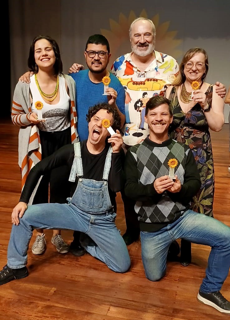 A imagem mostra sete pessoas em um palco de madeira, aparentemente após uma apresentação ou premiação. Todas elas estão sorrindo e segurando pequenos troféus em forma de girassóis. No centro, um homem mais velho com camisa colorida e barba branca está entre duas mulheres. Uma delas está à sua esquerda, vestindo uma blusa listrada e um colar de contas; a outra à sua direita, com um vestido floral escuro e um colar dourado. À esquerda do homem mais velho, um homem mais jovem com camisa azul está ao lado da mulher com a blusa listrada. À direita, um outro homem mais jovem com um suéter de losango está ao lado da mulher com o vestido floral. Na frente deles, dois homens mais jovens estão ajoelhados. Um deles usa um macacão jeans e está segurando um girassol na frente do olho; o outro está vestindo jeans e um suéter de argyle.