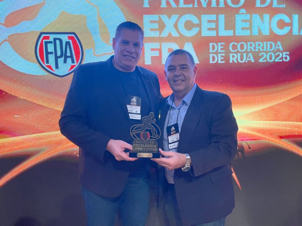 A imagem mostra dois homens em ternos escuros em pé na frente de um grande painel de fundo. O painel é principalmente laranja e amarelo, com o texto "Prêmio de Excelência de Corrida de Rua 2025" e um logotipo que parece dizer "FPA". Os homens parecem estar em um evento de premiação, pois um deles está segurando um troféu. O troféu é escuro e tem uma figura de um corredor em execução. O texto no troféu diz "Prêmio de Excelência FPA de Corrida de Rua". Os homens estão sorrindo e parecem felizes por estarem juntos. O homem à esquerda está um pouco mais alto que o homem à direita. Ambos os homens parecem ter cabelos escuros e estão vestindo ternos escuros. O homem à esquerda está usando um crachá de identificação que parece dizer "Fernando Silva". O homem à direita está usando um crachá que parece dizer "Alexandre". A imagem é bem iluminada e a qualidade é boa. A imagem provavelmente foi tirada em um evento de premiação ou conferência.