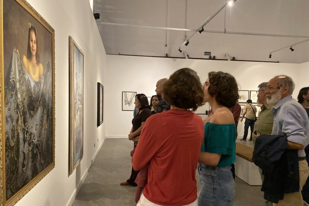 A imagem mostra uma galeria de arte com várias pessoas observando as obras de arte expostas. As obras incluem retratos e outras pinturas. As pessoas estão em grupos pequenos, conversando e apreciando as artes. A galeria é bem iluminada e espaçosa, com paredes brancas e pisos de concreto. Há uma saída visível no fundo da galeria. A atmosfera é calma e respeitosa, adequada para um ambiente de apreciação artística. Uma das pinturas mais proeminentes é um retrato de uma jovem em um vestido elaborado. O quadro é grande e está em uma moldura dourada. As outras pinturas são menores e parecem ser de estilos diferentes. As pessoas na imagem parecem ser visitantes da galeria, possivelmente participando de uma exposição ou visita guiada.