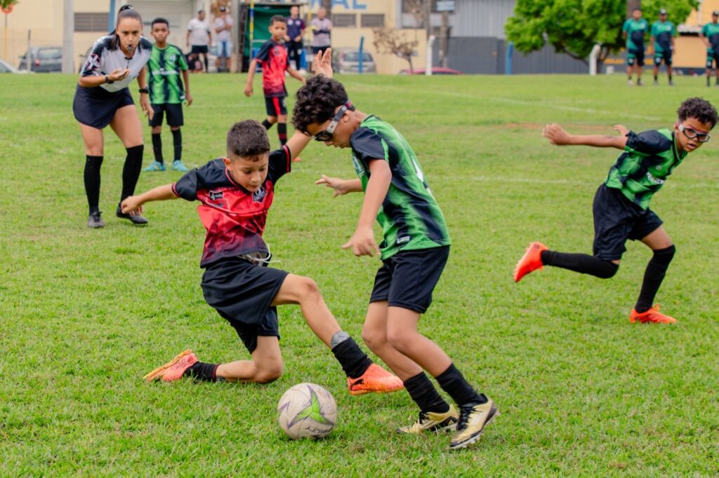 A imagem mostra uma partida de futebol infantil. No centro da imagem, dois meninos estão disputando a posse de bola, com um tentando roubar a bola do outro. Há uma árbitra mulher ao fundo, observando a partida. Outros jogadores estão visíveis ao fundo, tanto no campo quanto nas laterais. O campo é gramado e parece estar em boas condições. Um terceiro menino corre ao fundo, também envolvido na partida. A imagem captura a dinâmica e a energia de uma partida de futebol juvenil.