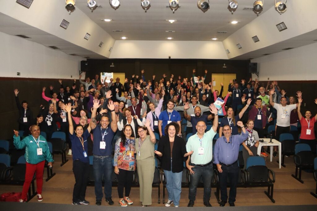 A imagem mostra uma grande multidão de pessoas em um auditório, todas com os braços erguidos. Elas parecem estar participando de um evento ou apresentação, e parecem felizes e entusiasmadas. A foto é tirada de um ângulo de nível do chão, mostrando o grupo inteiro. As pessoas estão sentadas em cadeiras, e há um palco no fundo. A imagem é bem iluminada, e as cores são vibrantes. A atmosfera geral é positiva e festiva.
