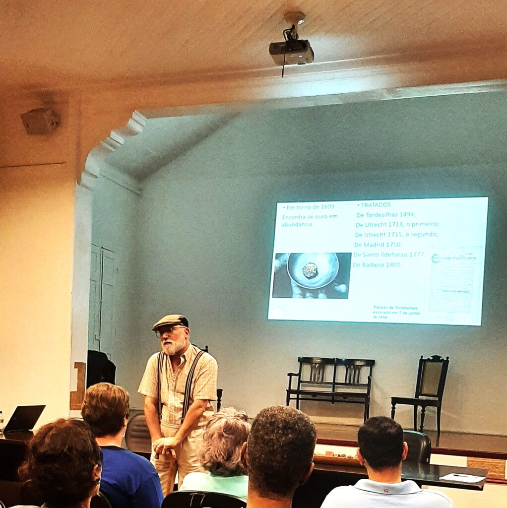 A imagem mostra uma apresentação numa sala de palestras. Um homem de meia-idade, com chapéu e barba grisalha, está de pé no centro da sala, aparentemente a dar uma palestra. A tela de projeção atrás dele apresenta informações sobre o ouro e os tratados históricos, especificamente mencionando os tratados de Tordesilhas, Utrecht e Madrid, com as respectivas datas. A sala é clássica, com cadeiras de madeira e uma atmosfera formal. A imagem sugere uma palestra ou aula sobre história, possivelmente relacionada à colonização e exploração de recursos na América do Sul. Há um público sentado na frente, ouvindo atentamente a apresentação.