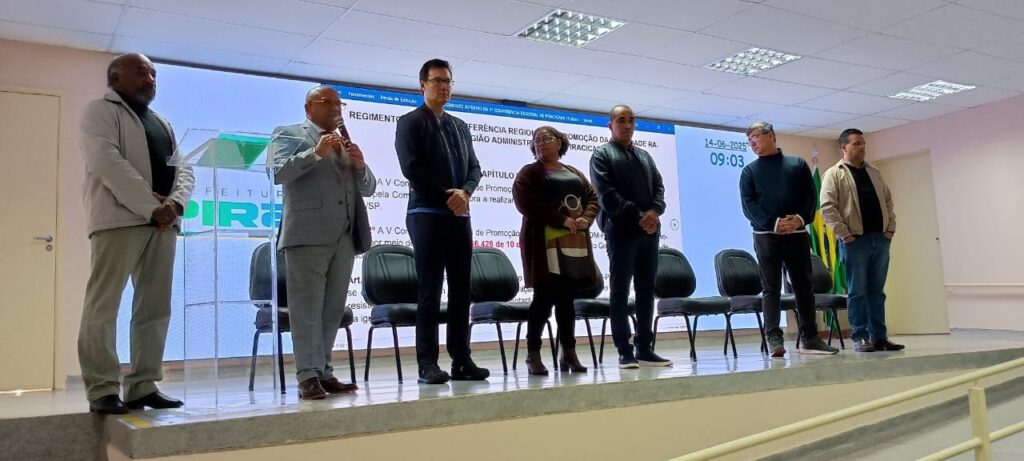 A imagem mostra uma sala de conferências onde um evento está ocorrendo. Há um palco com várias pessoas sentadas em cadeiras. Uma tela grande atrás deles exibe informações sobre a conferência, incluindo o nome, data e hora. A tela também mostra um logotipo que parece ser o da prefeitura de Piracicaba. As pessoas no palco parecem estar envolvidas na conferência ou apresentação. A sala parece limpa e organizada, e há uma atmosfera formal. No geral, a imagem parece ser um registro de uma conferência ou reunião formal.