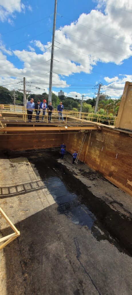 A imagem mostra um grupo de pessoas em uma plataforma alta observando o interior de um grande tanque retangular. O tanque parece ser de concreto ou metal, e seu interior está parcialmente cheio de um líquido escuro. Há duas escadas no tanque, uma de madeira e outra de metal. O céu está parcialmente nublado, com algumas nuvens brancas e fofas. Ao fundo, há árvores e postes de energia. As pessoas na plataforma parecem estar usando roupas de trabalho ou casuais. A cena parece ser um local industrial ou de construção. A imagem é bem iluminada e o contraste é bom, permitindo uma boa visualização de todos os detalhes.