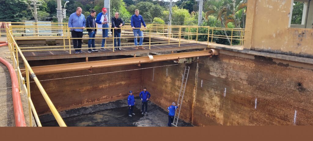 A imagem mostra uma vista aérea de uma estrutura de concreto, aparentemente uma estação de tratamento de água. Há uma piscina retangular no centro da imagem, com água escura e um fundo de concreto rachado. Uma escada de alumínio está encostada em uma das paredes da piscina. Na parte superior da estrutura, há uma passarela com corrimão amarelo, onde um grupo de pessoas, provavelmente funcionários ou inspetores, estão observando a piscina. Há também alguns trabalhadores na parte inferior da estrutura, perto da piscina. A área ao redor da piscina parece antiga e necessitar de manutenção. Há também uma mangueira laranja visível na parte inferior direita da imagem. O ambiente é externo, com vegetação visível ao fundo. O dia parece ensolarado.