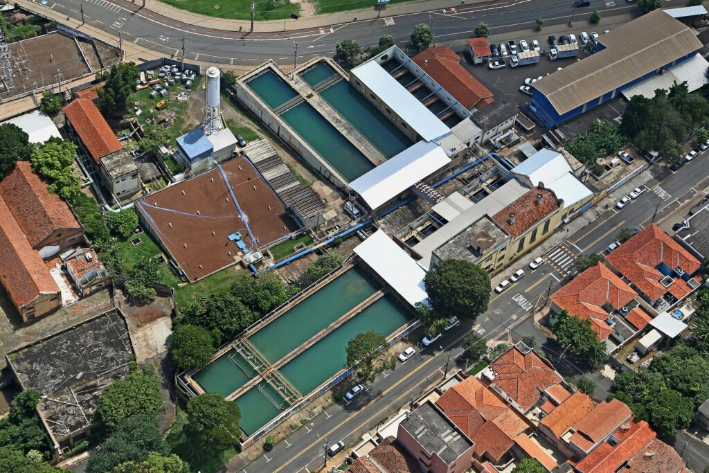 A imagem aérea mostra uma estação de tratamento de água, rodeada por edifícios residenciais e ruas. A estação possui tanques retangulares de água, tubulações e diversos edifícios auxiliares. As casas ao redor são principalmente de um andar, com telhados vermelhos. As ruas são pavimentadas e há carros estacionados ao longo delas. A vegetação é esparsa, com algumas árvores e arbustos visíveis. A imagem sugere uma área urbana de densidade média, com uma infraestrutura de tratamento de água presente. A perspectiva aérea permite uma visão abrangente da disposição dos edifícios e da infraestrutura.