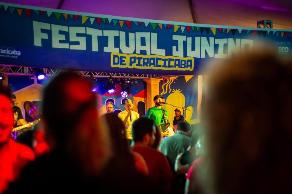 A imagem mostra uma cena vibrante e animada de um festival de música ao ar livre, especificamente o "Festival Junino de Piracicaba". Um grande banner azul vibrante com o nome do festival está pendurado acima do palco. O palco está decorado com motivos coloridos e alegres, com desenhos que lembram as festas juninas tradicionais. Uma banda está se apresentando no palco, com vários músicos visíveis. O vocalista principal se destaca usando uma camisa verde brilhante. Outros membros da banda estão usando roupas mais escuras. A multidão na frente do palco é parcialmente visível, com rostos e formas parcialmente obscurecidos, sugerindo uma atmosfera lotada e animada. As luzes do palco projetam cores vibrantes sobre a multidão, adicionando à atmosfera festiva. No geral, a imagem captura a energia e a alegria de um festival de música ao vivo, com foco no evento específico do Festival Junino de Piracicaba. A qualidade da foto é alta, e a composição captura bem a atmosfera do evento.