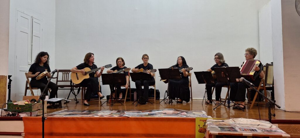 A imagem mostra um grupo de sete mulheres tocando música em um palco. Elas estão sentadas em cadeiras individuais, com microfones posicionados à frente. Cincom delas tocam violões clássicos, uma toca pandeiro e outra um acordeão. As mulheres usam roupas escuras e parecem estar em um ambiente formal, possivelmente um concerto ou apresentação musical. O fundo é uma parede branca simples, e o palco é de madeira. Há alguns objetos na frente do palco, que parecem ser parte da apresentação ou decoração do local. A imagem é bem iluminada e nítida, permitindo a visualização clara das mulheres e dos instrumentos. A atmosfera geral da imagem sugere um evento musical tranquilo e organizado.