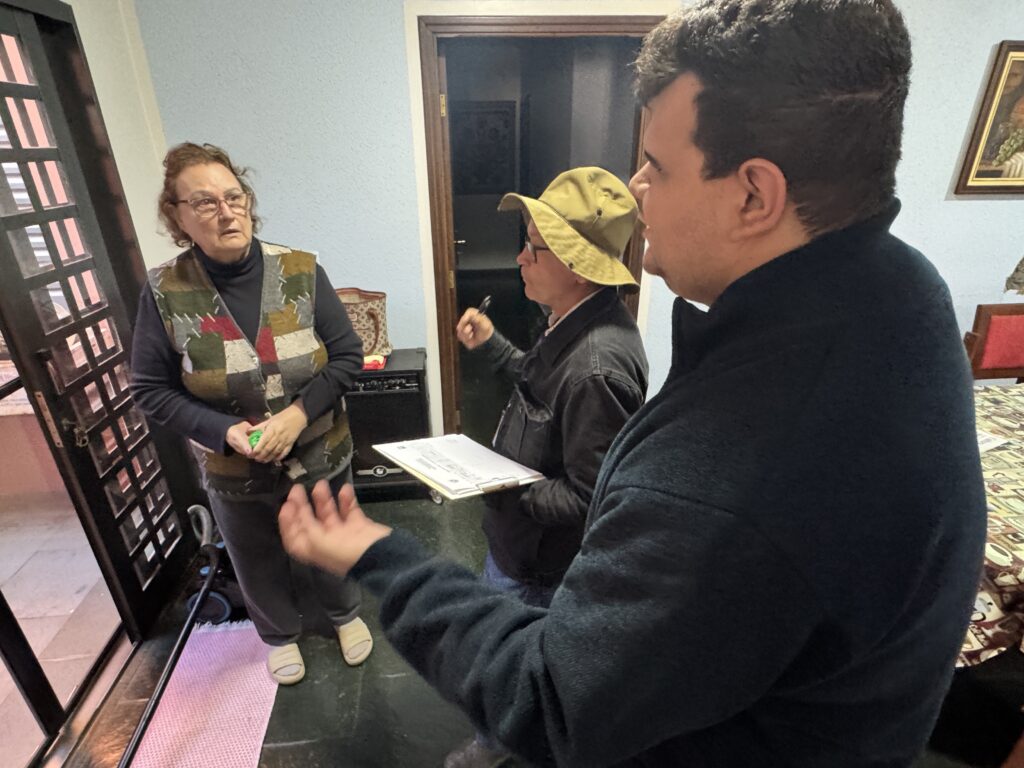 A imagem mostra três pessoas em uma sala. Uma mulher de meia-idade está de pé perto de uma porta, vestindo um colete acolchoado multicolorido sobre uma blusa escura e calças pretas. Ela segura algo pequeno nas mãos. Ao lado dela, está um homem mais jovem usando um chapéu bege e uma jaqueta escura. Ele segura uma prancheta com papéis. O terceiro indivíduo, um homem mais jovem com cabelo escuro, está de costas para a câmera, mas parece estar interagindo com as outras duas pessoas. Ele está vestindo uma jaqueta escura. A sala parece ser uma casa, com uma porta de madeira escura, uma mesa parcialmente visível e um amplificador musical no canto. A atmosfera é informal e parece ser uma conversa casual.