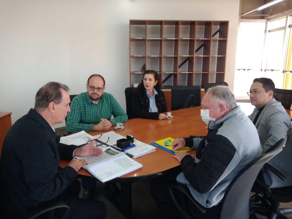 Cinco pessoas participam de uma reunião em torno de uma mesa oval, com documentos, carimbo e xícaras de café. O grupo discute pautas formais, em ambiente corporativo e organizado. Um dos presentes usa máscara, o que pode indicar preocupação com saúde ou protocolos.