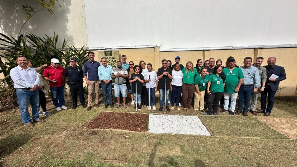 A imagem mostra um grande grupo de pessoas em pé em frente a um muro de tijolos. Há cerca de 30 pessoas na foto, incluindo homens e mulheres de diferentes idades e etnias. Algumas pessoas estão usando uniformes, enquanto outras estão usando roupas casuais. Algumas pessoas estão usando óculos de sol. Há dois pedaços de terra no chão na frente do grupo. Um pedaço de terra é coberto com lascas de madeira, enquanto o outro pedaço de terra é coberto com cascalho branco. No canto superior direito da imagem, há um banner que diz "Semana Municipal do Meio Ambiente". A imagem parece ter sido tirada em um evento ou reunião. As pessoas na imagem parecem estar felizes e engajadas. A imagem é bem composta e bem iluminada.