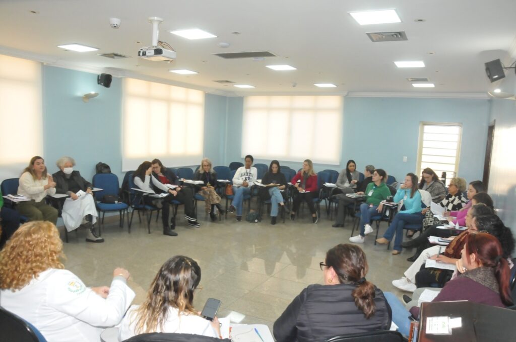 A imagem mostra um grupo de mulheres sentadas em cadeiras em círculo numa sala de reuniões. A sala é pintada de azul claro e tem janelas grandes com luz natural. Há um projetor no teto e as paredes são de cor azul claro. As mulheres parecem estar envolvidas numa discussão ou apresentação. Algumas delas estão a tomar notas e outras estão a olhar para a pessoa que está a falar. A atmosfera parece ser profissional e séria. As mulheres estão vestidas com roupas formais e informais.