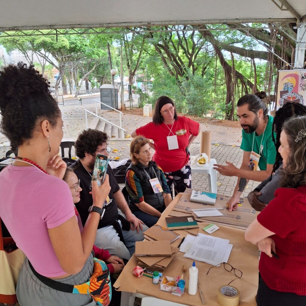 A imagem mostra um grupo de pessoas sentadas ao redor de uma mesa ao ar livre, aparentemente envolvidas em uma atividade artesanal ou de criação. A mesa está coberta com materiais como papelão, cadernos, cola, barbante e outras ferramentas de artesanato. As pessoas parecem estar colaborando em um projeto, com algumas pessoas olhando para o trabalho de outras e algumas tirando fotos com seus telefones. O ambiente é um local externo sombreado, com árvores e vegetação ao fundo. As pessoas na imagem parecem ser de diversas idades e origens, e todas estão vestidas de forma casual. Há um cartaz de fundo que sugere que a atividade pode ser parte de um evento ou feira. A atmosfera geral da imagem é descontraída e colaborativa.