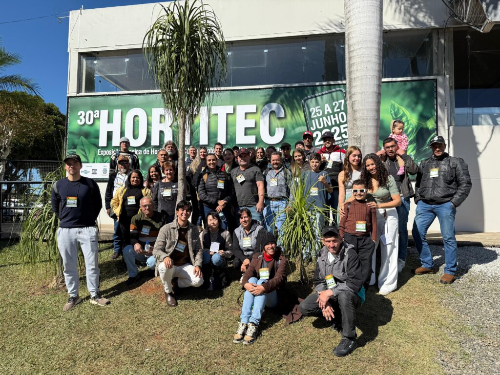 A imagem mostra um grande grupo de pessoas reunidas em frente a um banner que anuncia a "30ª Hortitec", uma exposição horticultura que ocorreu de 25 a 27 de junho de 2025. O grupo é composto por adultos e crianças, a maioria vestindo roupas casuais e crachás de identificação do evento. Eles estão sorrindo e posam para uma foto em grupo. Há uma árvore de palmeira alta ao lado do grupo, e o fundo é um prédio moderno com grandes janelas. A cena é tomada ao ar livre, em um dia ensolarado. O ambiente parece ser um local de evento ou feira. A imagem transmite uma atmosfera amigável e festiva, sugerindo uma reunião de participantes da Hortitec.