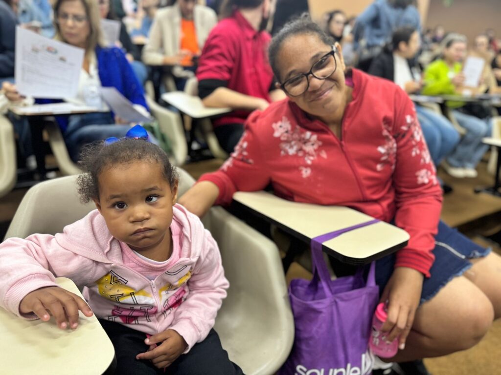 A imagem mostra uma mulher negra com óculos e uma jaqueta vermelha sentada em uma cadeira em uma sala de aula. Ela está segurando a mão de uma criança pequena de pele escura com uma jaqueta rosa. A criança está sentada em uma cadeira ao lado da mulher. Há outras pessoas sentadas em cadeiras ao redor delas, mas estão fora de foco. A sala de aula parece estar cheia de pessoas. A mulher e a criança parecem estar confortáveis ​​e felizes. A mulher está sorrindo para a câmera. A criança está olhando para a câmera com uma expressão séria. A mulher está usando uma sacola roxa. Há mesas na frente de cada cadeira.