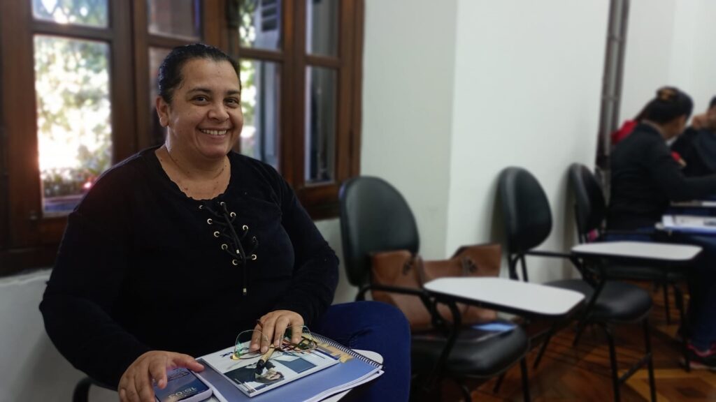 A imagem mostra uma mulher de meia-idade com pele morena sorrindo para a câmera. Ela está sentada em uma cadeira em uma sala de aula, com outras cadeiras e pessoas ao fundo levemente desfocadas. A mulher veste uma blusa preta de mangas compridas com um detalhe de amarração no pescoço e calças azuis escuras. Ela segura um caderno e óculos na mão. O ambiente parece ser uma sala de aula ou sala de estudos, com mesas individuais e cadeiras pretas. A iluminação é suave e natural, provavelmente vindo de uma janela grande visível à esquerda da imagem. A janela mostra árvores e folhagem ao fundo, sugerindo um ambiente externo arborizado. As outras pessoas no fundo estão sentadas em suas mesas, mas estão bastante fora de foco, tornando a mulher o centro principal da imagem. A imagem sugere um ambiente acadêmico ou de aprendizado.