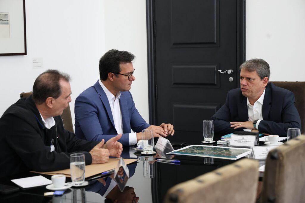 A imagem mostra três homens sentados em torno de uma mesa de conferência escura e brilhante. A mesa é polida e reflete os homens e os objetos sobre ela. Há documentos, canecas de café, copos de água e canetas espalhados pela mesa. Um mapa ou plano parece estar no centro da mesa, aberto e visível. O homem à esquerda da imagem está sentado com a postura ereta, usando uma jaqueta escura e uma camisa de gola alta por baixo. Ele parece estar envolvido em uma conversa séria, com as mãos juntas e um olhar concentrado. Seu cabelo é curto e escuro. O homem no centro está usando um blazer azul e uma camisa branca. Ele usa óculos e tem uma expressão séria, quase pensativa. Seus braços estão cruzados na frente dele, e ele parece estar prestando atenção ao que está sendo discutido.