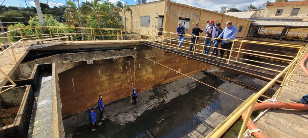 A imagem mostra um grupo de pessoas inspecionando uma estrutura de concreto grande e retangular que parece ser um tanque de armazenamento ou parte de um sistema de tratamento de água. Há dois grupos de pessoas: um grupo de seis homens está em uma plataforma elevada com uma grade de proteção, observando o tanque; outro grupo de três homens está no fundo do tanque, aparentemente realizando alguma inspeção ou manutenção. O tanque parece estar parcialmente cheio de um líquido escuro e turvo. As paredes do tanque mostram sinais de desgaste e deterioração, com manchas e marcas visíveis no concreto.