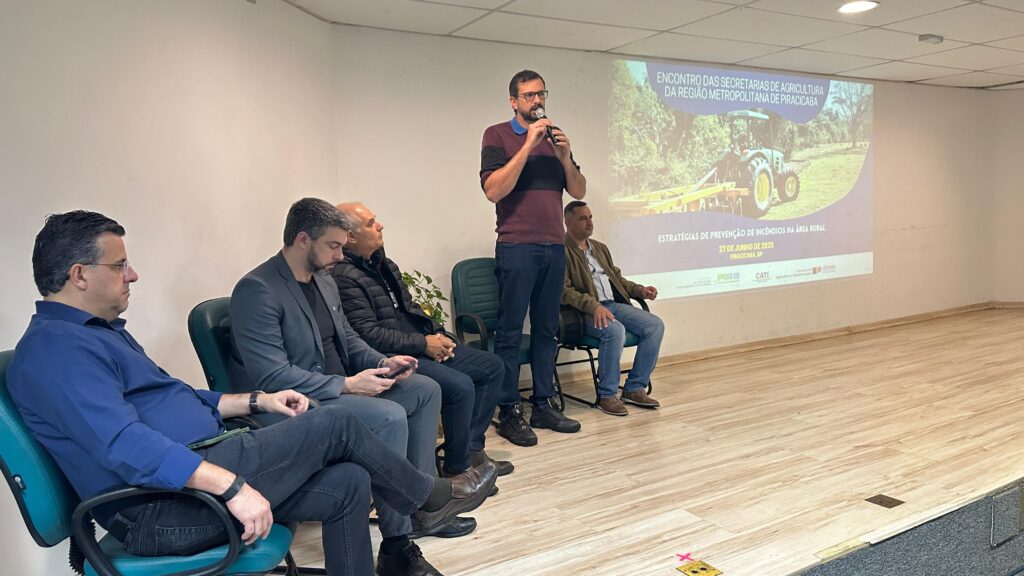A imagem mostra um painel de discussão em uma sala de palestras. Cinco homens estão sentados em cadeiras, um deles está de pé usando um microfone e falando. Há uma apresentação de slides na parede, mostrando o título "Encontro das Secretarias de Agricultura da Região Metropolitana de Piracicaba" e "Estratégias de Prevenção de Incêndios na Área Rural". Os homens parecem estar envolvidos em uma discussão sobre agricultura e prevenção de incêndios. O ambiente é formal e profissional. A sala tem um piso de madeira clara e cadeiras verdes.