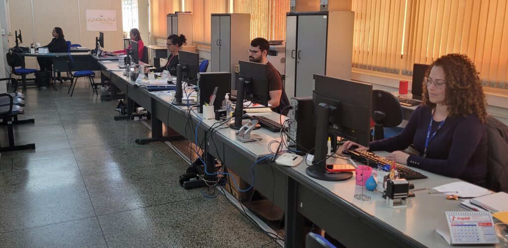 A imagem mostra uma sala de escritório com várias pessoas trabalhando em seus computadores. As mesas são longas e compartilhadas, com vários computadores e equipamentos de escritório. Há um ambiente de trabalho organizado, com cada pessoa em seu espaço individual. As pessoas parecem estar concentradas em suas tarefas. A iluminação é clara e natural, vindo de uma janela com persianas. O chão é de cerâmica clara, e as paredes são claras também. O ambiente é simples, mas funcional. Há arquivos e outros materiais de escritório sobre as mesas. No geral, a imagem transmite a ideia de um ambiente de trabalho eficiente e produtivo.