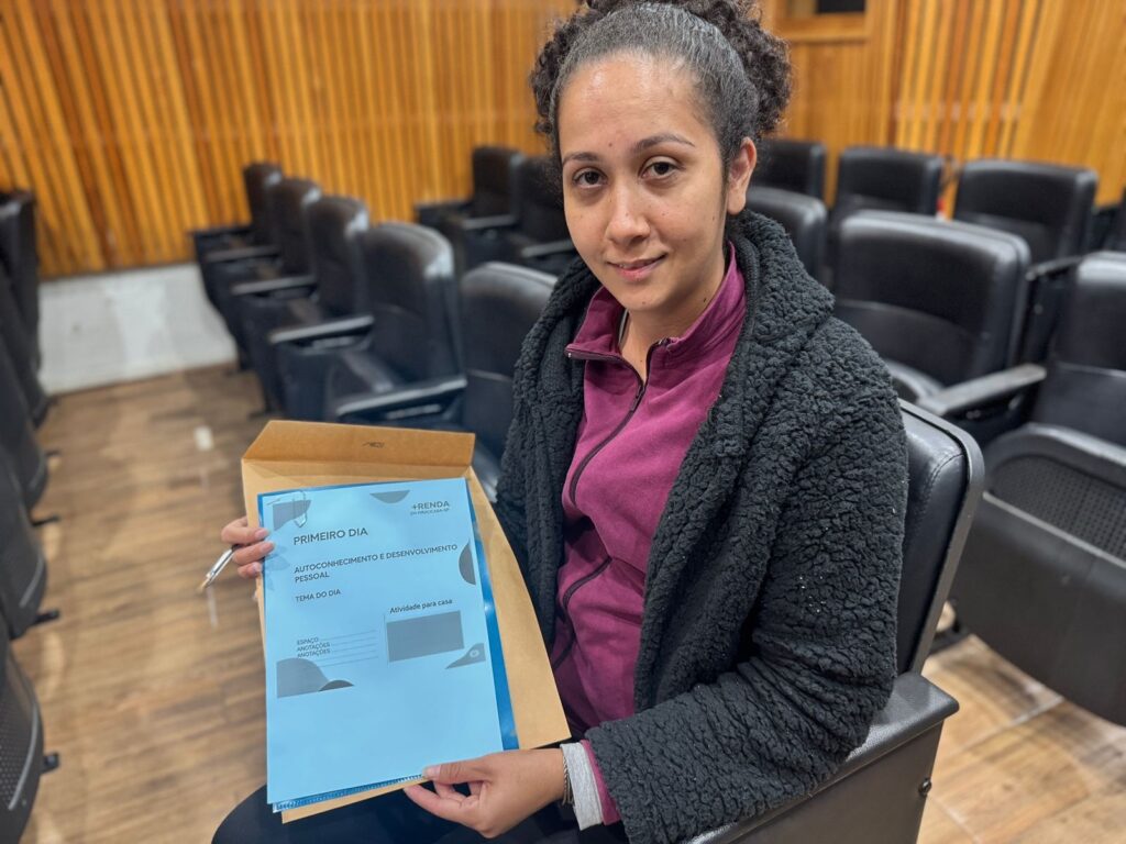 A imagem mostra uma jovem mulher sentada em uma cadeira em uma sala de aula ou auditório. Ela está segurando um documento em uma pasta parda. O documento é um material de um evento chamado "ARENDA em Piracicaba-SP", e é intitulado "Primeiro Dia - Autoconhecimento e Desenvolvimento Pessoal". A mulher parece calma e atenta, e está vestida com uma blusa roxa e uma jaqueta preta felpuda. O fundo da imagem mostra várias cadeiras vazias dispostas em fileiras, indicando um ambiente formal de aprendizado ou reunião. A iluminação é suave, e a imagem tem uma qualidade geral clara e nítida. A expressão facial da mulher sugere concentração ou expectativa.